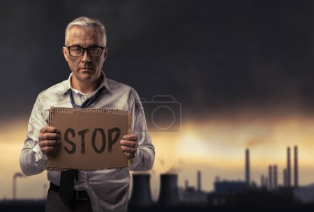 Foto de Sucio empresario desesperado sosteniendo un cartel y protestando contra la contaminación, la planta industrial y los gases tóxicos en el fondo - Imagen libre de derechos