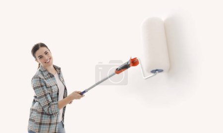 Foto de Mujer feliz pintando paredes en su nuevo hogar, ella está usando un rodillo de pintura, pancarta con espacio para copiar - Imagen libre de derechos