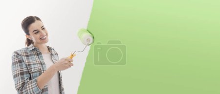 Foto de Joven mujer sonriente renovando y decorando su nueva casa, ella está usando un rodillo de pintura y aplicando pintura verde a las paredes - Imagen libre de derechos