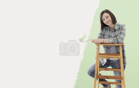 Foto de Feliz joven pintando paredes en su nuevo hogar, ella está sosteniendo un rodillo de pintura y sonriendo, espacio de copia - Imagen libre de derechos
