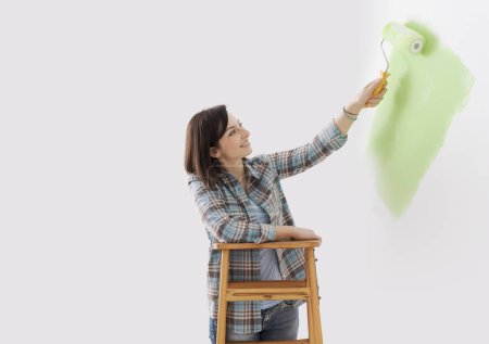 Téléchargez les photos : Heureuse jeune femme peignant des murs dans sa nouvelle maison, elle tient un rouleau de peinture et sourit, espace de copie - en image libre de droit