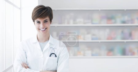 Photo for Young professional pharmacist posing with arms crossed and smiling, store interior in the background - Royalty Free Image