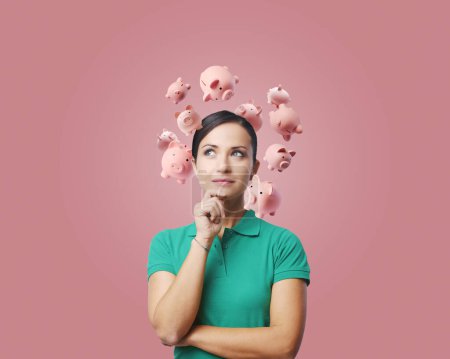 Photo for Smart young woman thinking with hand on chin, she has many piggy banks around her head, financial planning and earning concept - Royalty Free Image