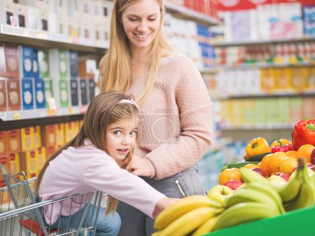 Foto de Feliz linda chica sentada en un carrito de compras y la compra de verduras y frutas frescas con su madre en el supermercado - Imagen libre de derechos