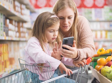 Téléchargez les photos : Jolie fille assise sur un panier d'achat au supermarché, sa mère montre ses applications sur le smartphone : concept de technologie et d'épicerie - en image libre de droit