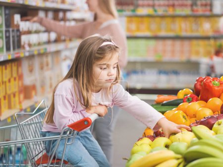 Photo for Cute lovely girl sitting on the shopping cart, she is taking a fruit in the produce section while her mother is busy taking products on the shelves in the background - Royalty Free Image