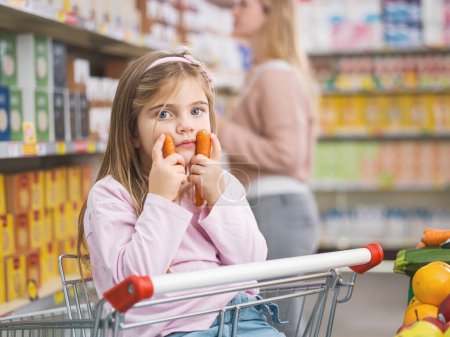 Photo for Portrait of a cute girl sitting on a shopping cart at the supermarket, she is holding carrots - Royalty Free Image