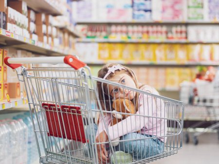 Téléchargez les photos : Petite fille mignonne au supermarché, elle est assise à l'intérieur d'un chariot et étreint son ours en peluche - en image libre de droit