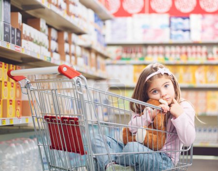 Portrait of a cute little girl at the supermarket, she is sitting inside a shopping cart and hugging her teddy bear