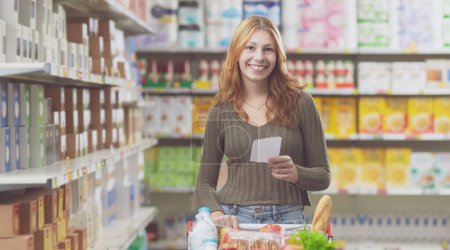 Téléchargez les photos : Jeune femme souriante faisant des courses au supermarché, elle pousse un chariot plein et tient une liste de courses - en image libre de droit