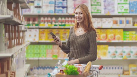 Téléchargez les photos : Portrait d'une jeune femme faisant de l'épicerie au supermarché, elle sourit et tient son smartphone - en image libre de droit