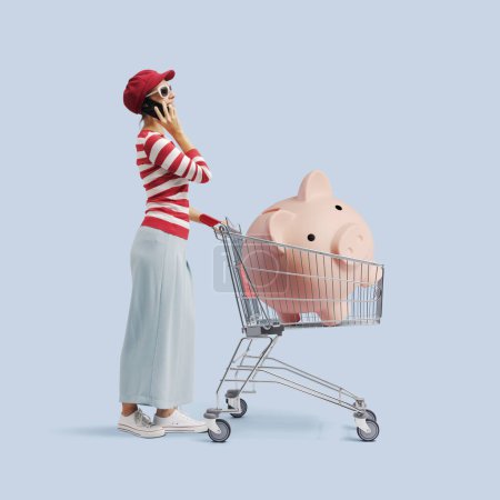 Photo for Happy woman having a phone call and pushing a trolley containing a big piggy bank, shopping on a budget concept - Royalty Free Image