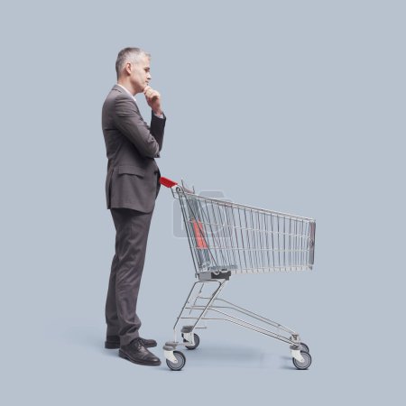 Foto de Pensativo dudoso hombre de negocios de pie detrás de un carrito de compras, él está pensando con la mano en la barbilla - Imagen libre de derechos