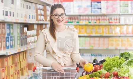 Foto de Mujer sonriente empujando un carrito de la compra y la compra de verduras frescas y saludables en el supermercado, compras de comestibles y concepto de nutrición - Imagen libre de derechos