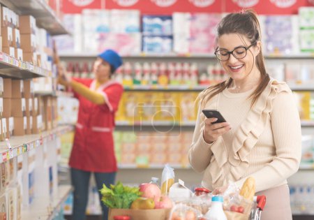 Téléchargez les photos : Femme souriante achetant des produits d'épicerie et utilisant un smartphone à l'épicerie - en image libre de droit