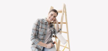 Téléchargez les photos : Jeune femme souriante peignant sa maison, elle se tient à côté d'une échelle et tient un rouleau de peinture - en image libre de droit