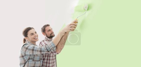 Foto de Joven pareja amorosa renovando su casa, están pintando paredes y sosteniendo un rodillo de pintura juntos - Imagen libre de derechos