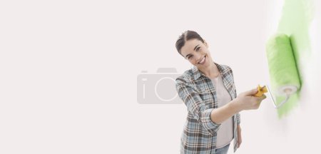 Téléchargez les photos : Heureuse femme peignant des murs dans sa nouvelle maison, elle utilise un rouleau de peinture, bannière avec espace de copie - en image libre de droit