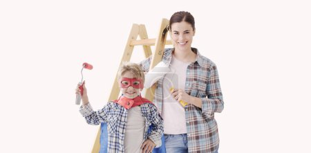 Téléchargez les photos : Joyeux jeune mère et garçon souriant peignant leur maison, l'enfant porte un costume de super héros - en image libre de droit