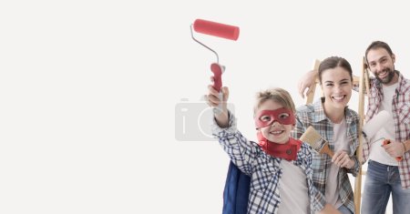 Foto de Familia feliz renovando su casa, están pintando paredes y sonriendo a la cámara, el niño lleva un disfraz de superhéroe - Imagen libre de derechos
