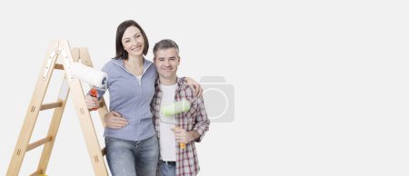 Téléchargez les photos : Heureux jeune couple rénovant leur maison et leurs salles de peinture, ils posent avec des rouleaux de peinture et sourient - en image libre de droit