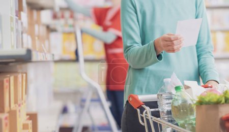 Foto de Cliente compra comestibles en el supermercado, ella está sosteniendo una lista de supermercados y empujando un carrito de compras completo - Imagen libre de derechos
