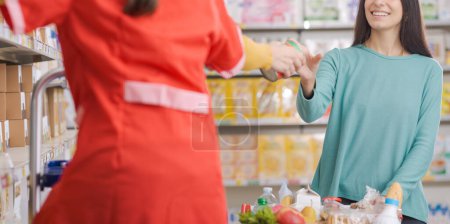 Téléchargez les photos : Commis à l'épicerie aider un client au supermarché, elle prend un produit de l'étagère et le lui donne - en image libre de droit