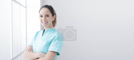Téléchargez les photos : Jeune infirmière confiante qui travaille à l'hôpital, elle pose les bras croisés et sourit - en image libre de droit