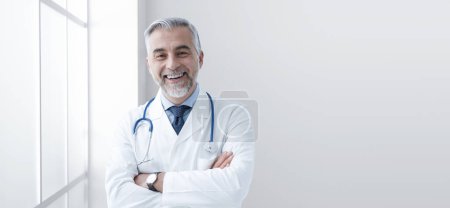 Foto de Médico de confianza sonriendo y posando con los brazos cruzados en el hospital, concepto de asistencia médica y sanitaria, espacio para copiar - Imagen libre de derechos