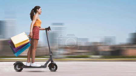 Foto de Mujer joven y feliz llevando muchas bolsas de compras y montando un scooter eléctrico, paisaje urbano en el fondo, venta y concepto de compras - Imagen libre de derechos
