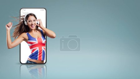 Téléchargez les photos : Adolescente souriante portant un débardeur drapeau anglais, elle écoute de la musique avec des écouteurs et danse dans un vidéocall smartphone - en image libre de droit