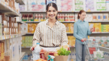 Foto de Joven hermosa mujer empujando un carrito de compras completo y la compra de comestibles en el supermercado - Imagen libre de derechos