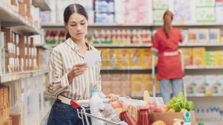 Foto de Joven mujer pensativa con la cesta de la compra completa en el supermercado, ella está revisando su lista de compras, concepto de compras de comestibles - Imagen libre de derechos