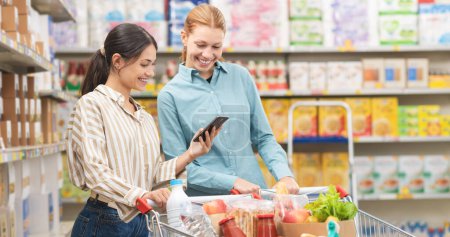 Photo for Smiling women at the supermarket buying groceries together, one woman is showing her smartphone to her friend, grocery shopping concept - Royalty Free Image