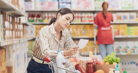 Téléchargez les photos : Jeune femme chère avec panier complet au supermarché, elle vérifie sa liste d'achats, concept d'épicerie - en image libre de droit