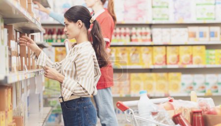 Téléchargez les photos : Jeune femme faisant des courses au supermarché, elle prend des produits de l'étagère du magasin et tient une liste d'achats - en image libre de droit