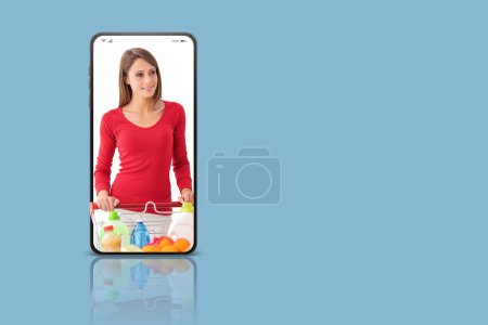 Foto de Aplicación de compras en línea: mujer sonriente empujando un carrito de compras completo en la pantalla de un teléfono inteligente - Imagen libre de derechos