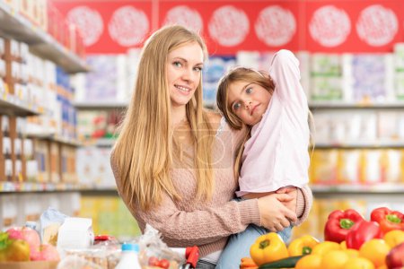 Foto de Feliz joven madre sosteniendo a su hija y haciendo compras en el supermercado, están sonriendo a la cámara - Imagen libre de derechos