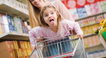 Téléchargez les photos : Fille heureuse faisant des courses avec sa mère, elle s'appuie sur le panier et sort sa langue - en image libre de droit