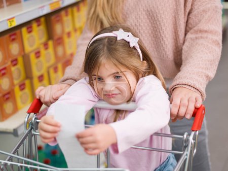 Téléchargez les photos : Fille déçue au supermarché avec sa mère, elle tient un long reçu d'épicerie coûteux - en image libre de droit