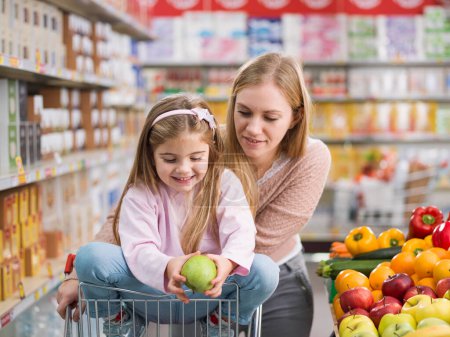 Foto de Feliz linda chica sentada en un carrito de compras y la compra de verduras y frutas frescas con su madre en el supermercado - Imagen libre de derechos
