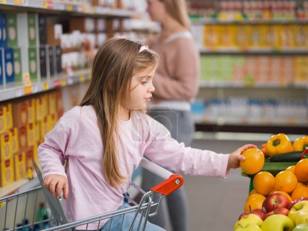 Foto de Chica inteligente sentada en el carrito de la compra y tomando frutas frescas y saludables en el supermercado, su madre está de pie en el fondo - Imagen libre de derechos