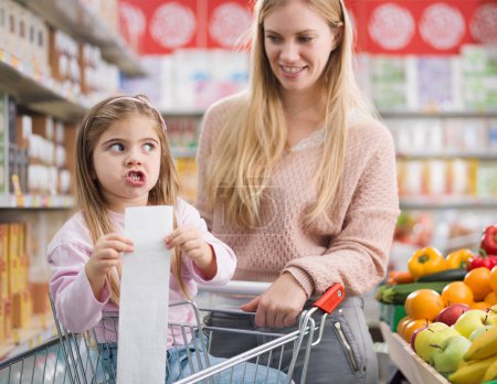 Enttäuschtes Mädchen im Supermarkt mit ihrer Mutter, sie hält einen teuren Einkaufszettel in der Hand