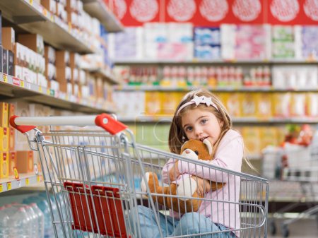 Foto de Linda chica sonriente en el supermercado, ella está sentada en un carrito de compras y abrazando a su oso de peluche - Imagen libre de derechos