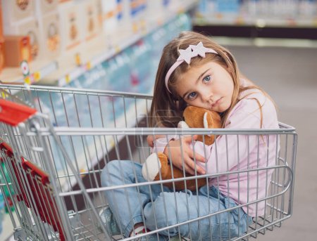 Retrato de una linda niña en el supermercado, ella está sentada dentro de un carrito de compras y abrazando a su osito de peluche