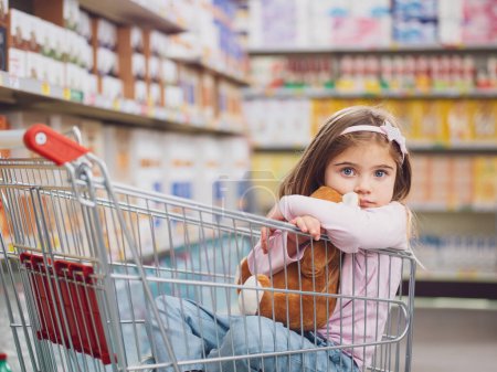 Porträt eines süßen kleinen Mädchens im Supermarkt, sie sitzt in einem Einkaufswagen und umarmt ihren Teddybär