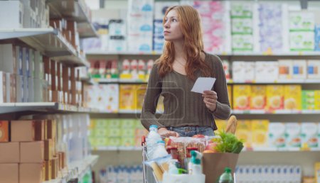 Foto de Mujer joven haciendo compras en el supermercado, ella está empujando un carro lleno y sosteniendo una lista de la compra - Imagen libre de derechos