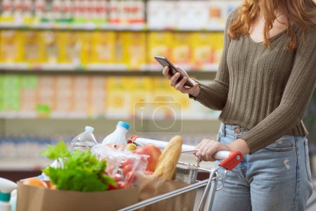 Foto de Mujer joven haciendo compras de comestibles en el supermercado, ella está empujando un carrito de compras completo y utilizando su teléfono inteligente - Imagen libre de derechos