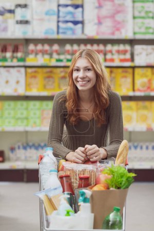 Photo for Portrait of a young woman at the supermarket, she is leaning on the shopping cart handle and smiling at camera - Royalty Free Image
