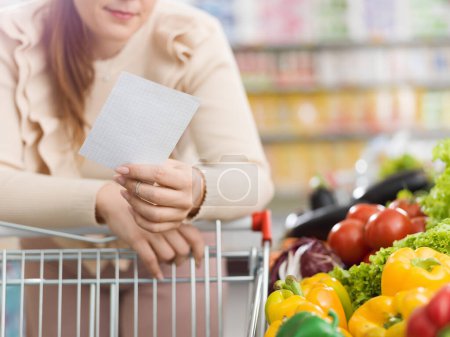 Téléchargez les photos : Femme faisant des courses au supermarché dans la section des produits, elle s'appuie sur le panier et tient une liste d'épicerie - en image libre de droit